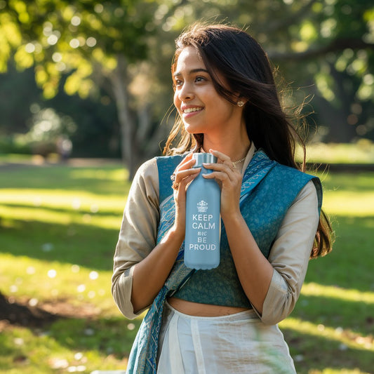 A young woman in a traditional outfit holding a light blue insulated water bottle with the words "Keep Calm and Be Proud" printed on it, smiling outdoors in a park.